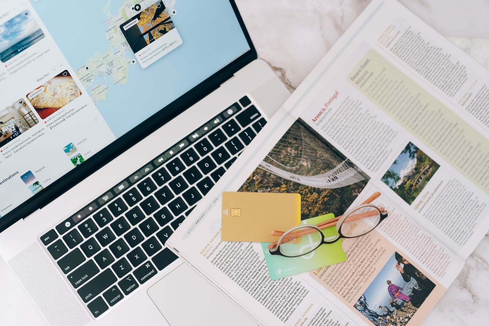 A laptop, travel brochure, glasses and a payment card on a desk
